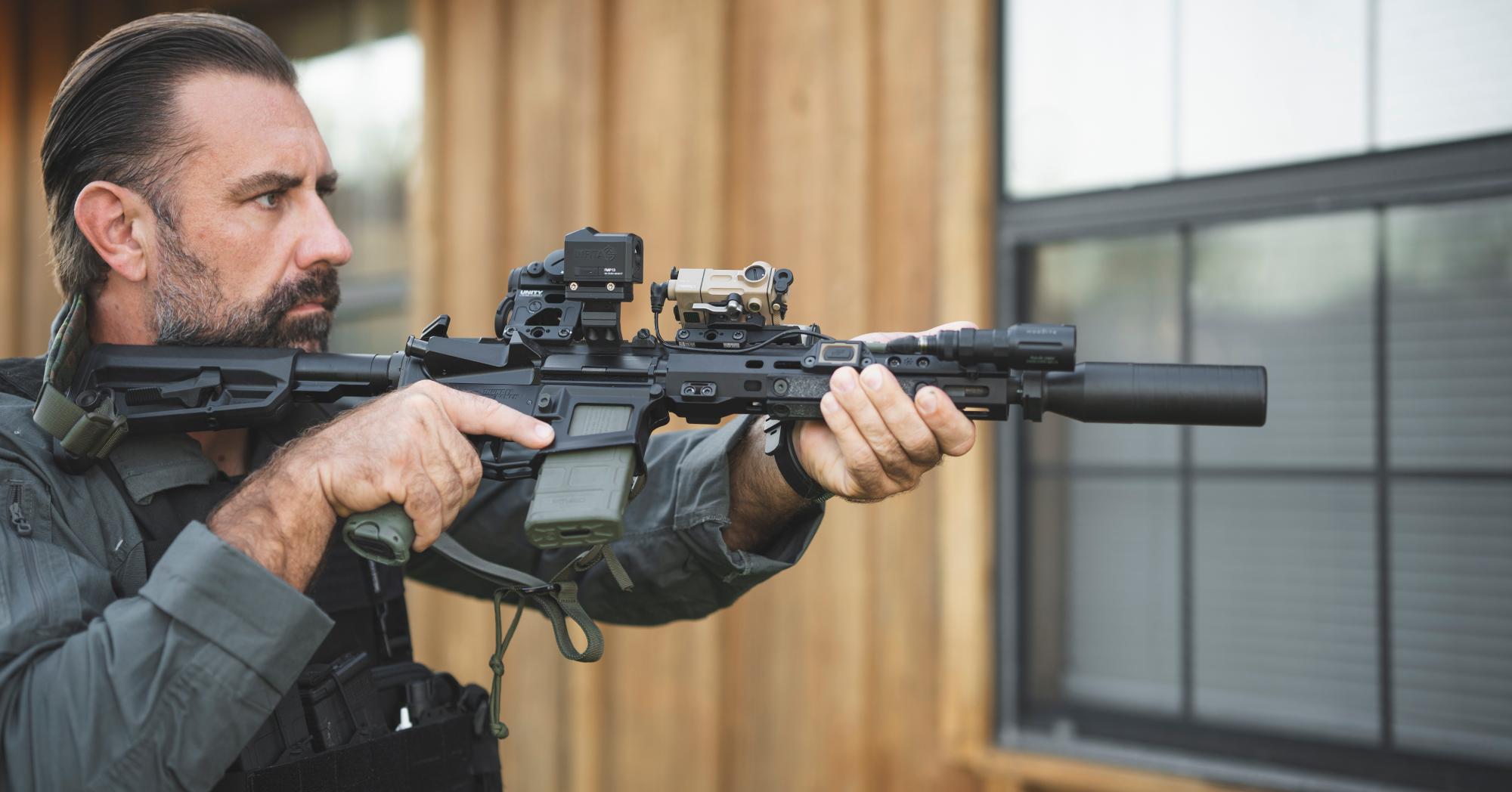 Side profile of a man in tactical gear aiming a modern black rifle equipped with a suppressor, flashlight, and multiple optics.