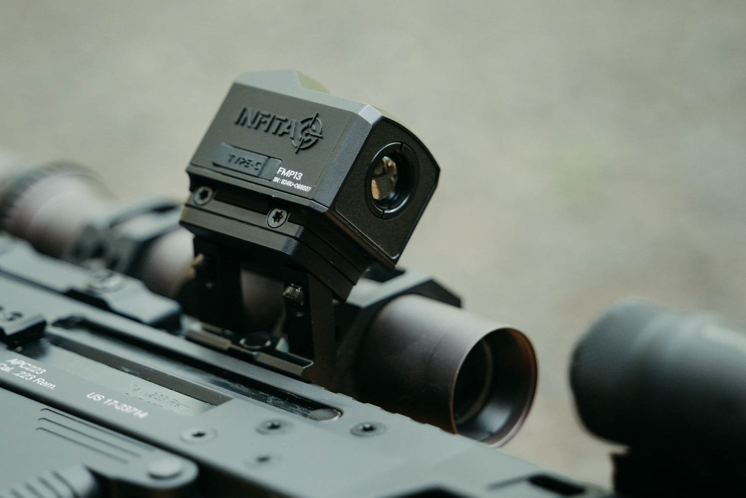 A close-up of a person holding a black rifle outfitted with a red dot sight and a tan laser module.