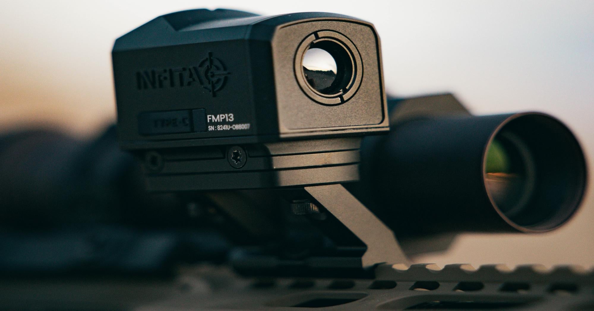 A close-up, outdoor shot of a black Infitac FMP13 thermal sight. The reflection of a distant, hazy landscape is visible in the sight's optic.