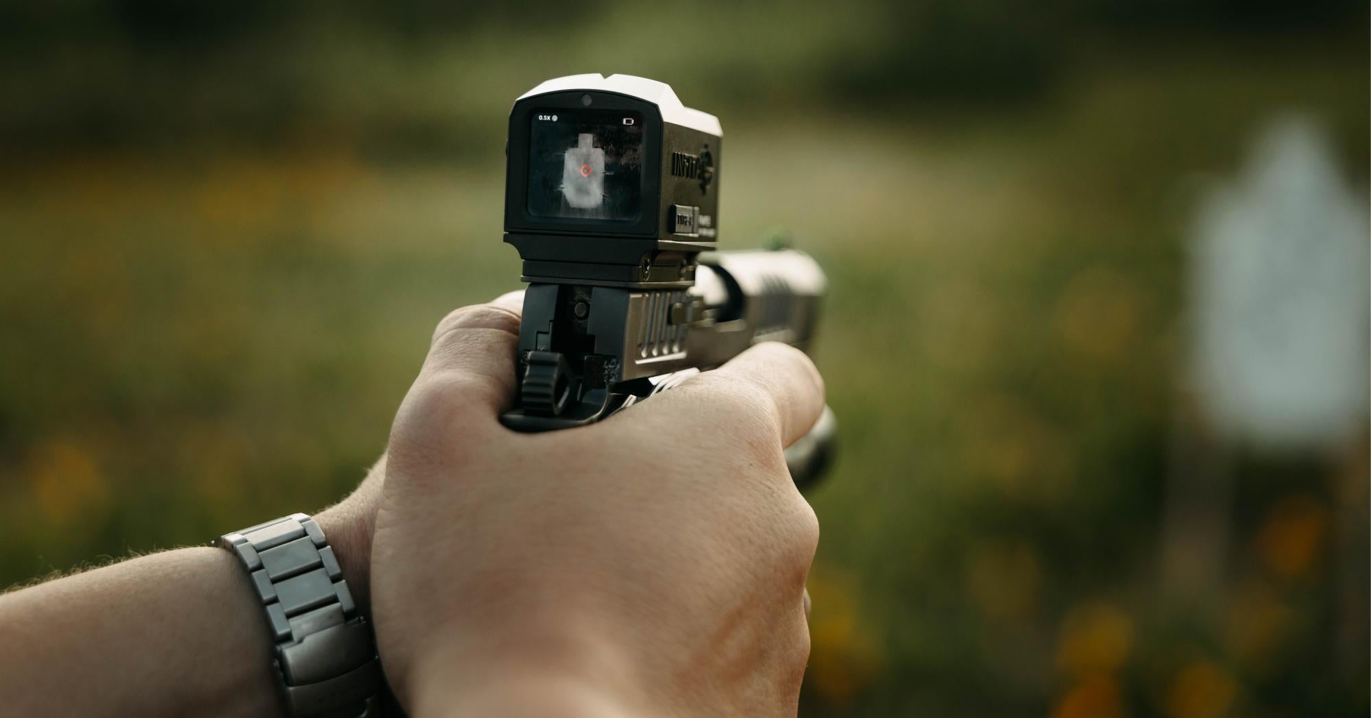 A first-person perspective of a shooter aiming a handgun equipped with a digital smart sight, which displays a clear image of the target on its screen.