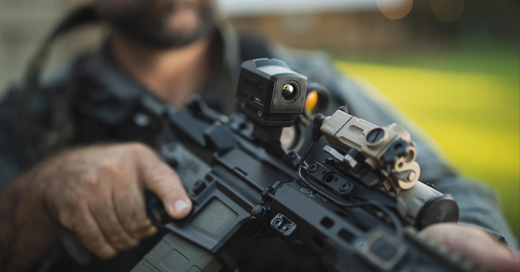 A close-up of a person holding a black rifle outfitted with a red dot sight and a tan laser module.