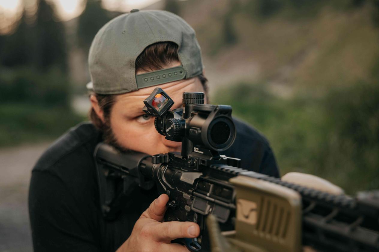 Shooter aims AR-style rifle with mounted red dot optic, green cap, outdoor setting
